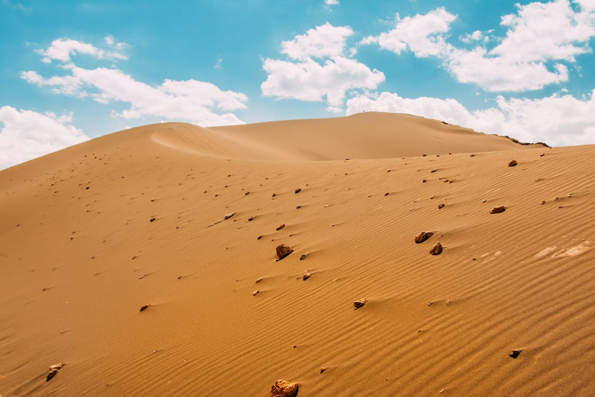 Golden sand dunes and scattered rocks beneath a turquoise sky and white clouds.