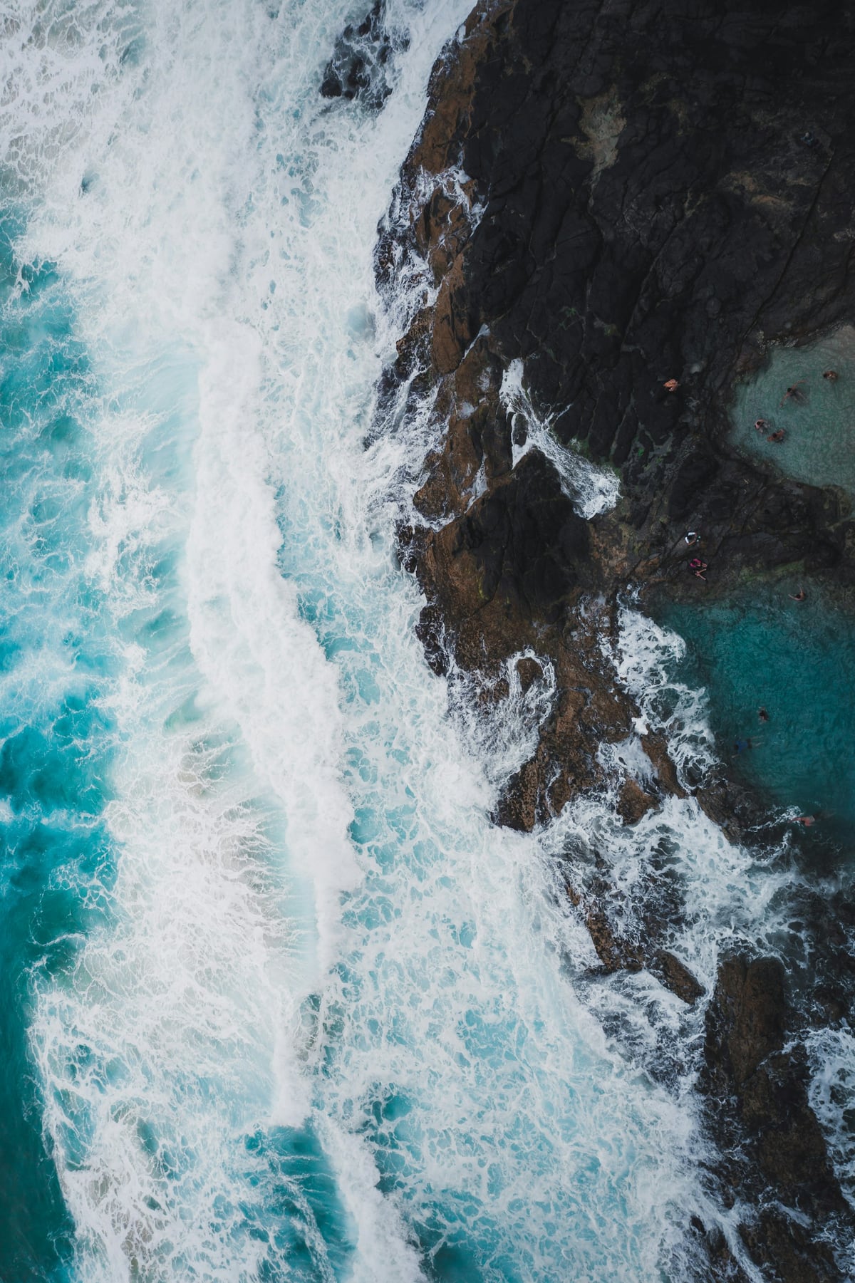 White surf breaks against dark jagged rocks in an aerial view of the coast.