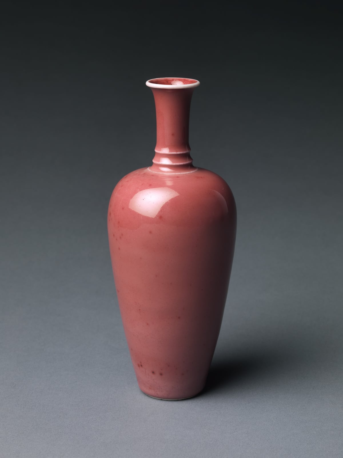 A slender pink-red glazed porcelain vase against a charcoal background.