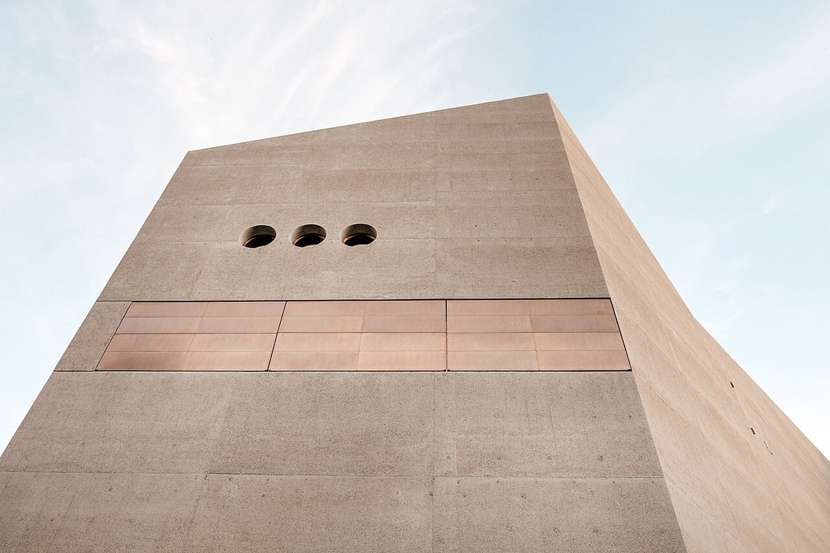 Pale concrete planes and three circular openings rise against a soft blue sky.